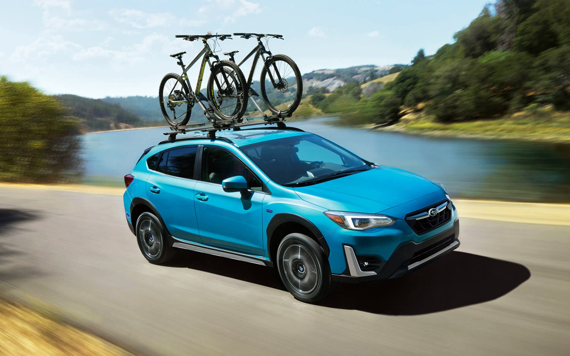 A blue Crosstrek Hybrid with two bicycles on its roof rack driving beside a river | Subaru of Ann Arbor in Ann Arbor MI